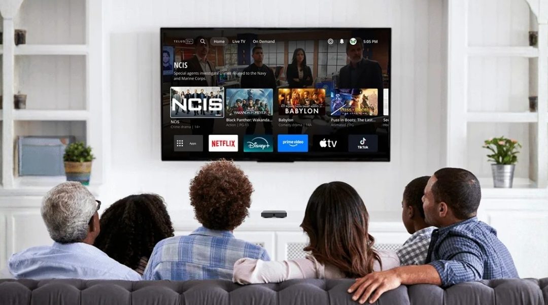Six people sit on a sofa watching a wall-mounted TV displaying a streaming app grid with icons for Netflix, Disney+, Prime Video and Apple TV.
